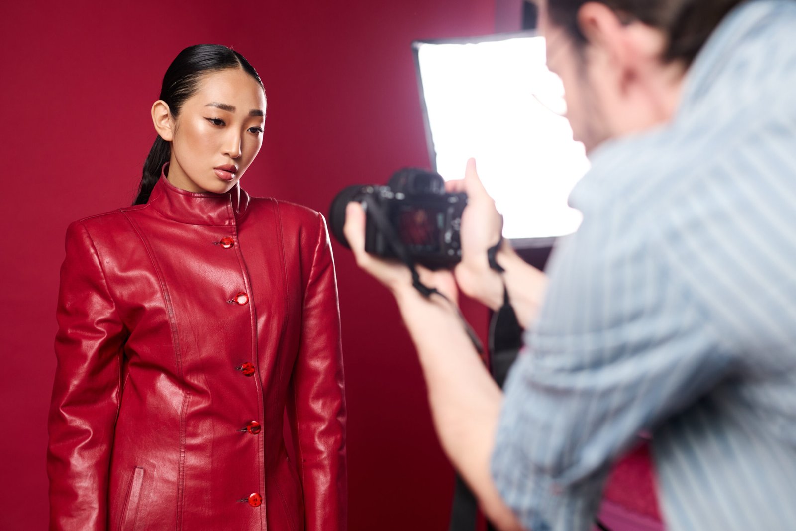 Asian brunette woman in red leather jacket posing for a photo with a man taking a picture. red background and light add a dramatic effect to the image.