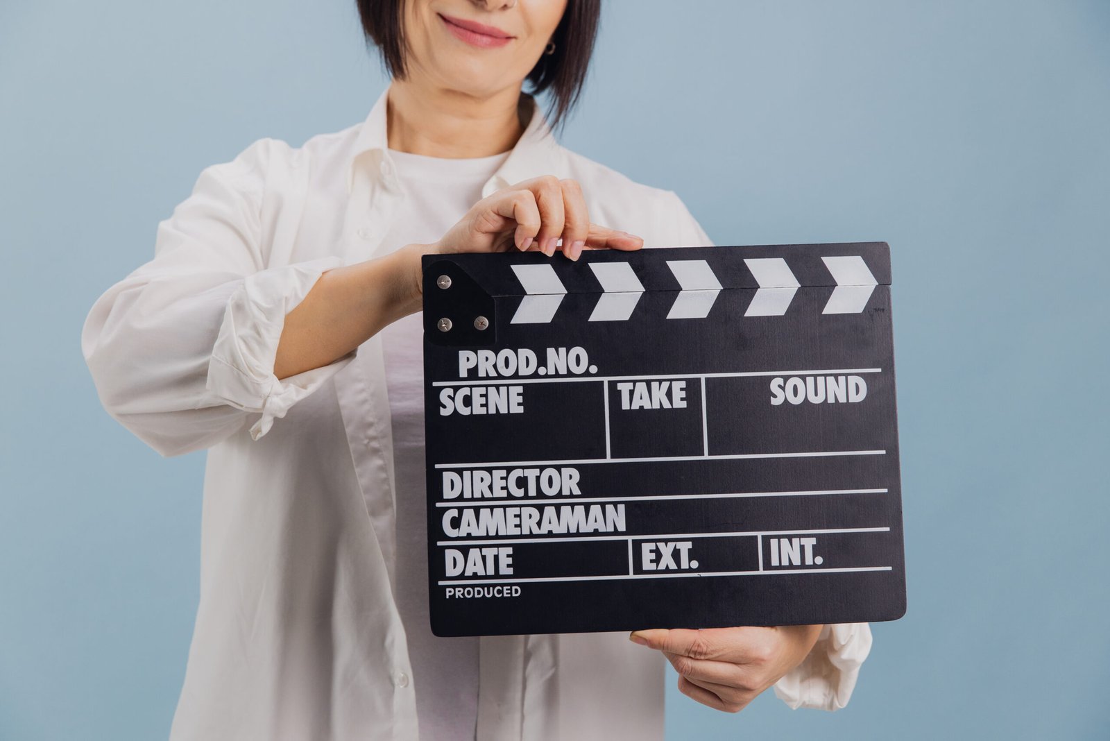 Smiling mature woman in white shirt holding a film clapperboard on blue background, ready to start shooting